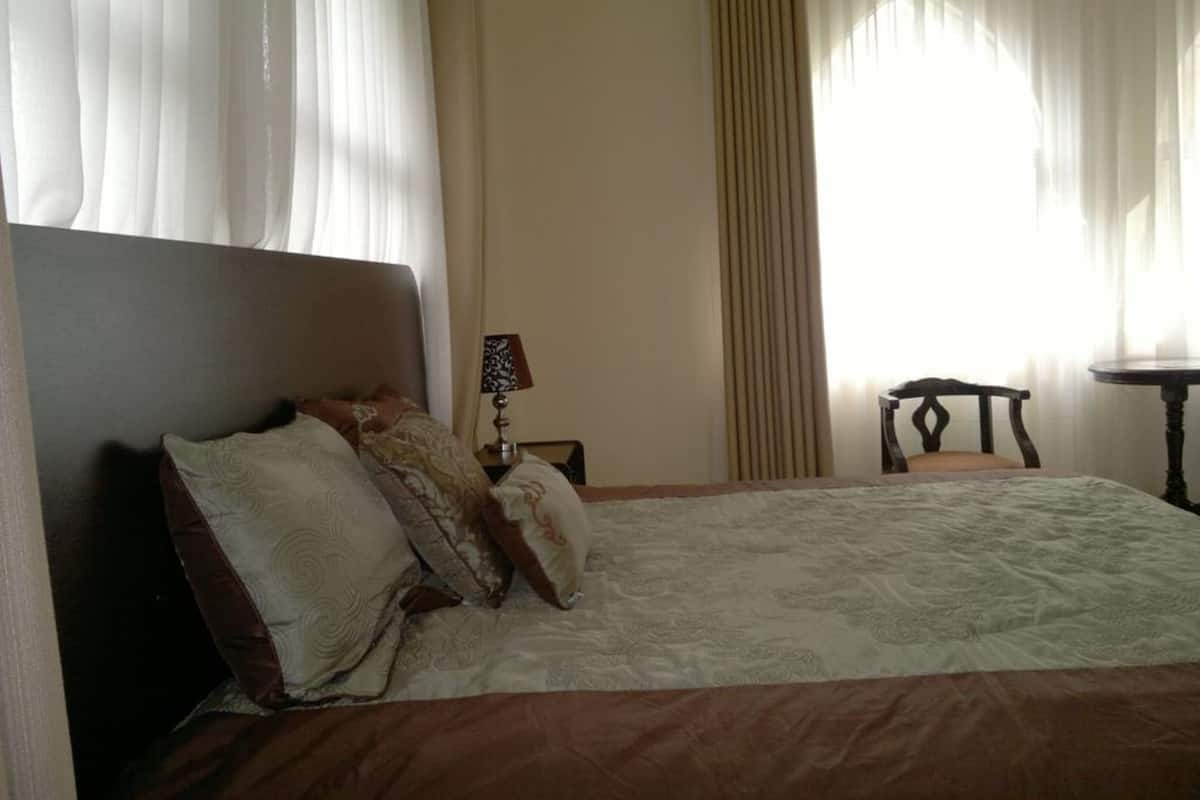 Une chambre à coucher avec un lit, une table de chevet, une lampe, une chaise et une fenêtre avec des rideaux.