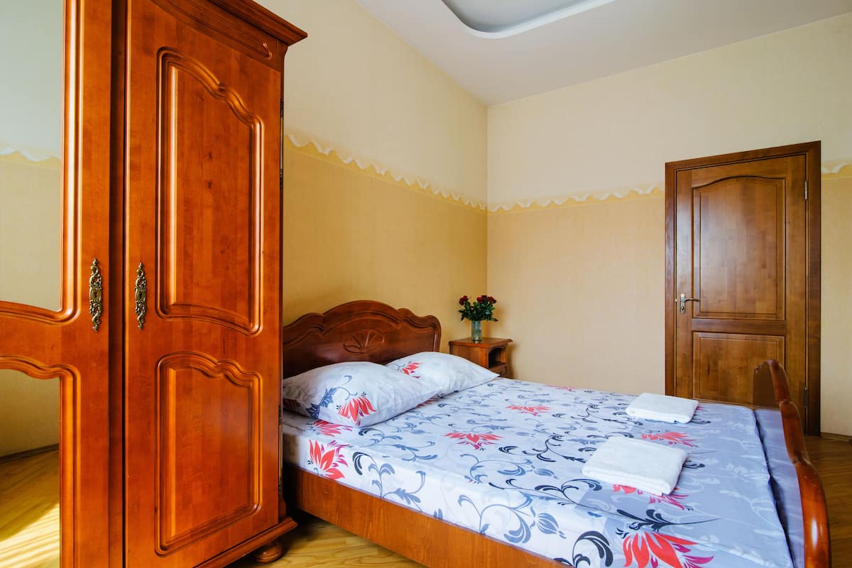A bedroom with a wooden wardrobe, a bed with floral bedding, and a nightstand with a vase.