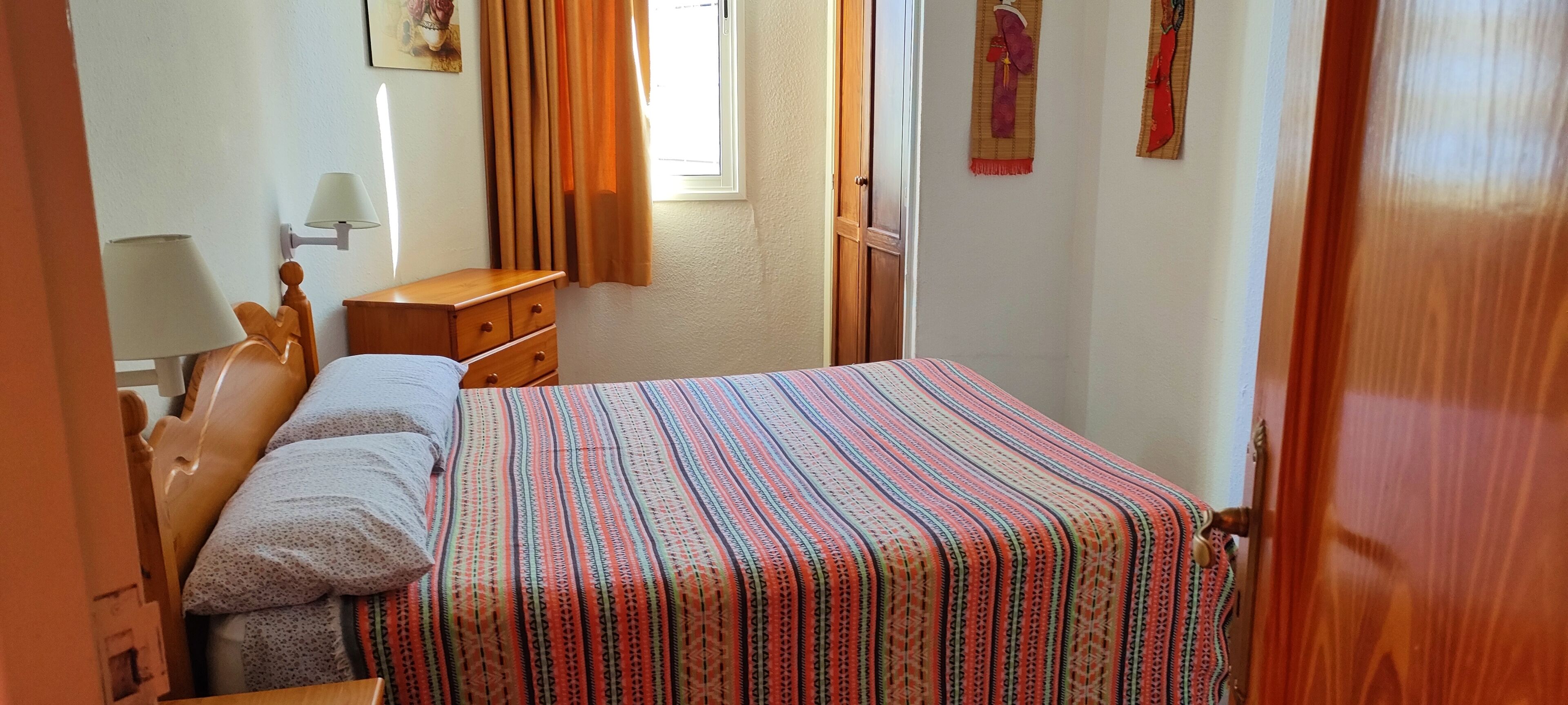 A bedroom with a bed, wooden headboard, a nightstand, a lamp, a window with curtains, and wall decorations.