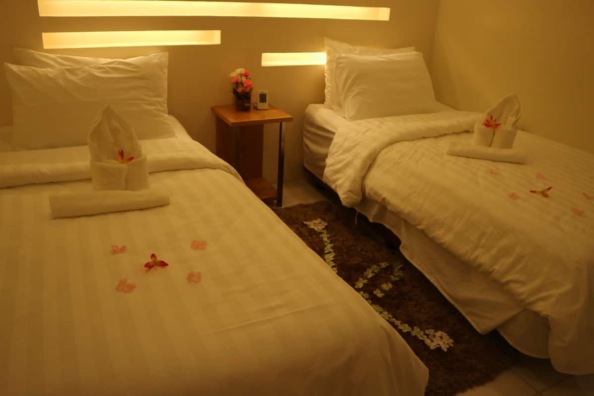 Two single beds with white linens, a small bedside table with a vase of flowers, and a brown rug with floral patterns.