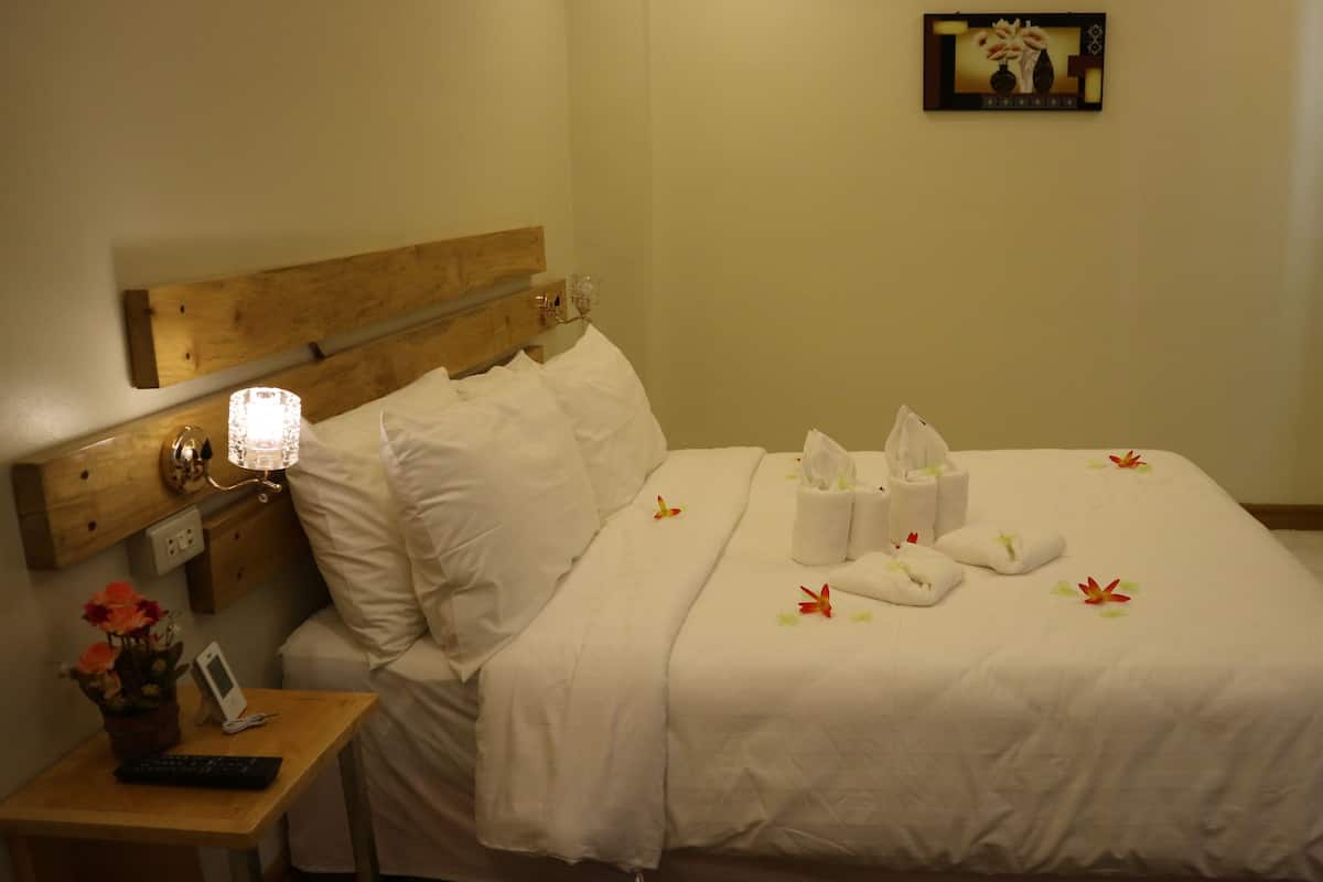 A neatly made bed with white linens, a bedside table with a remote control and a vase of flowers, and a wall-mounted picture frame.