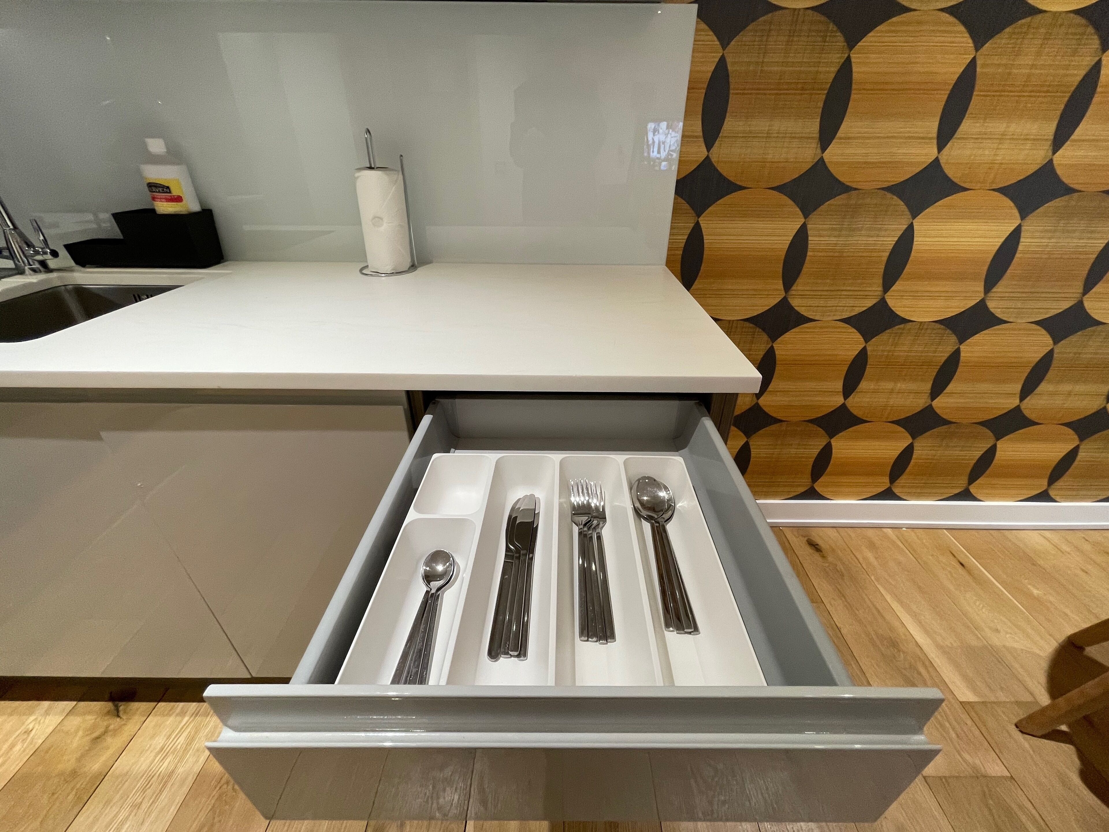 A kitchen with a white countertop, a wooden patterned wall, and a drawer containing utensils.