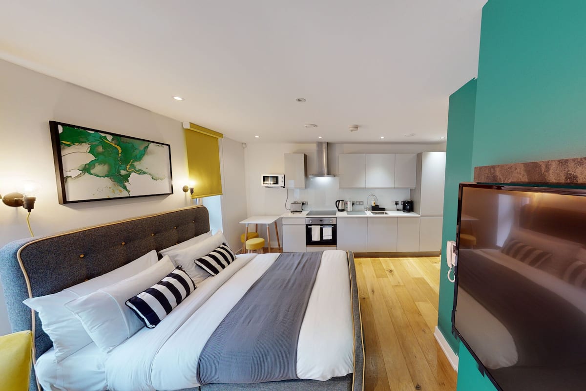 A modern hotel room with a bed, a TV, a kitchenette, and a dining area.