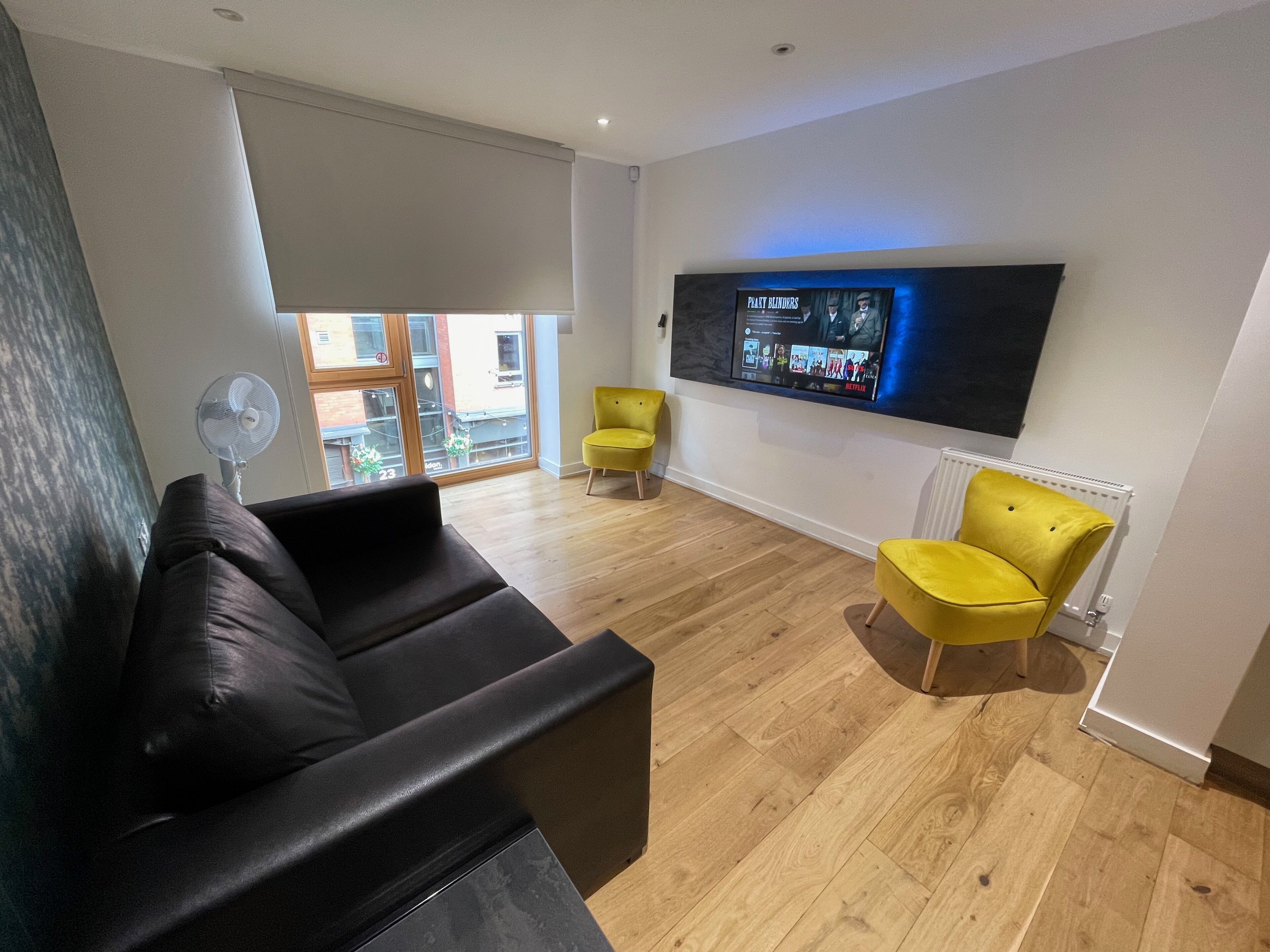 A modern living room with a black sofa, yellow chairs, a flat-screen TV, and a large window with a view of buildings.