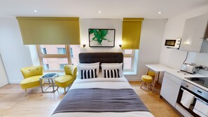 Ein modernes Hotelzimmer mit einem Bett, zwei gelben Sesseln, einem kleinen Tisch, einer Küchenzeile und einem großen Fenster mit Blick auf die Stadt.