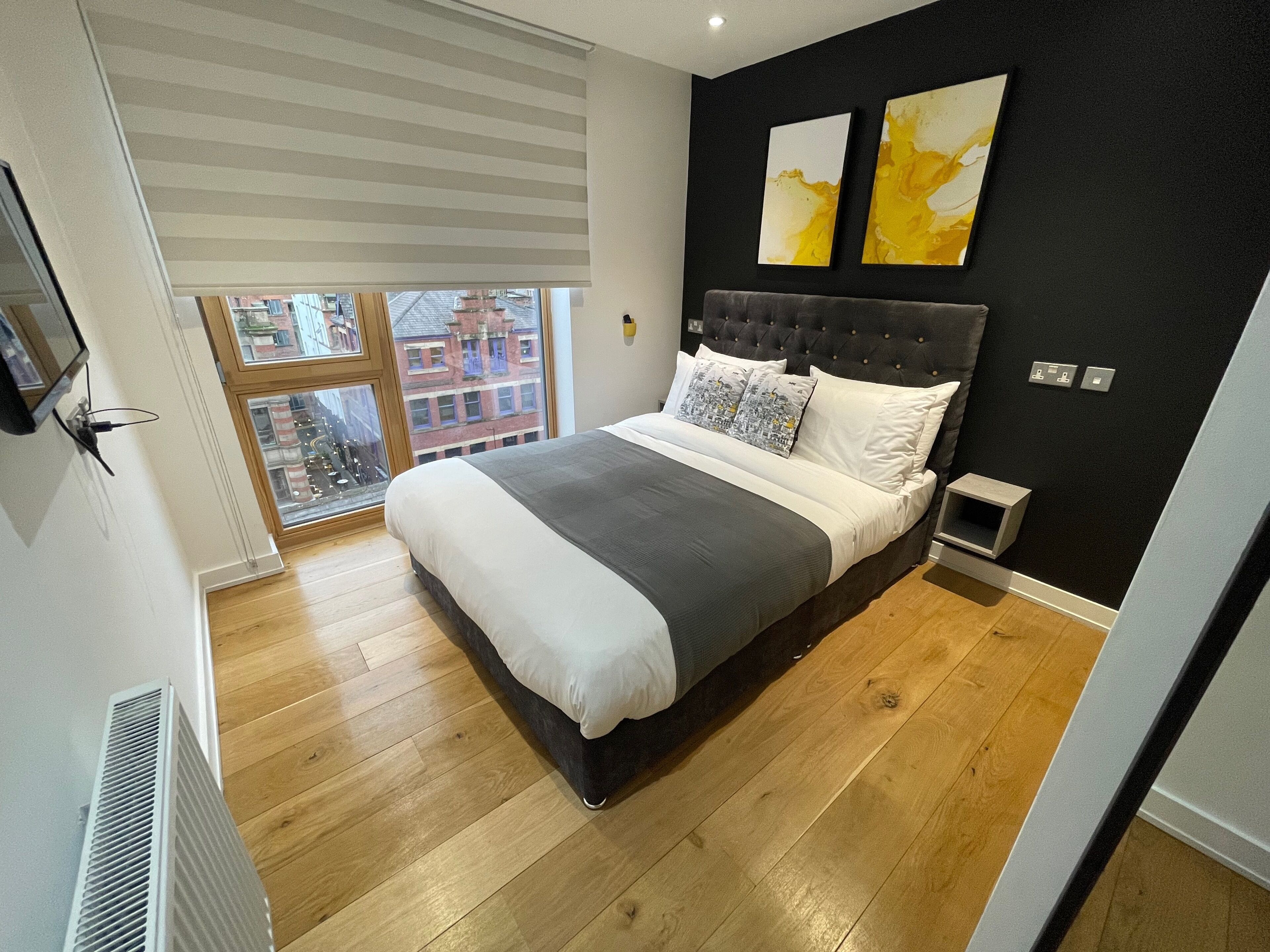 A modern bedroom with a large bed, a view of the city through a window, and two abstract paintings on the wall.