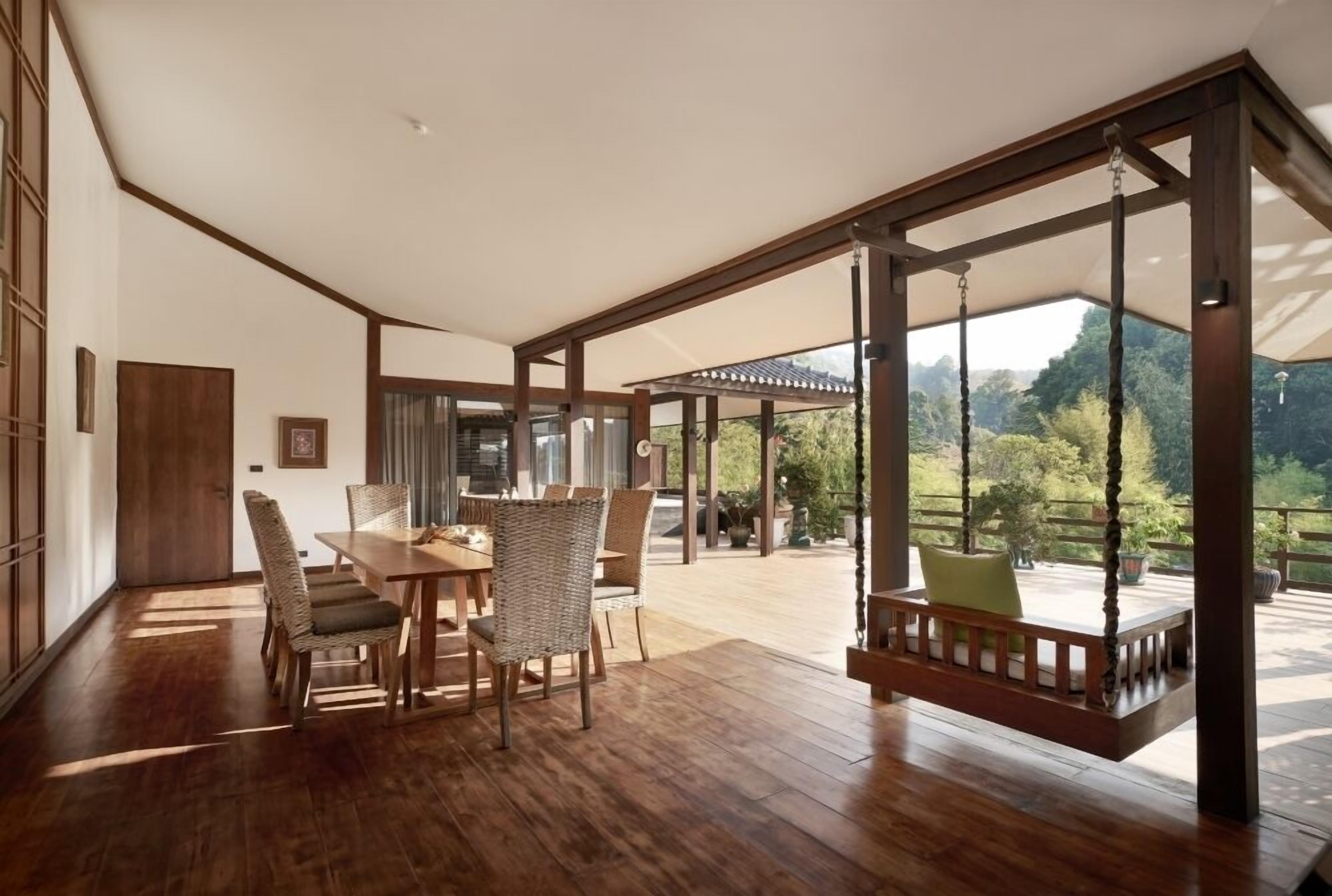 A spacious room with a wooden dining table, wicker chairs, a swing, and a view of a lush garden.