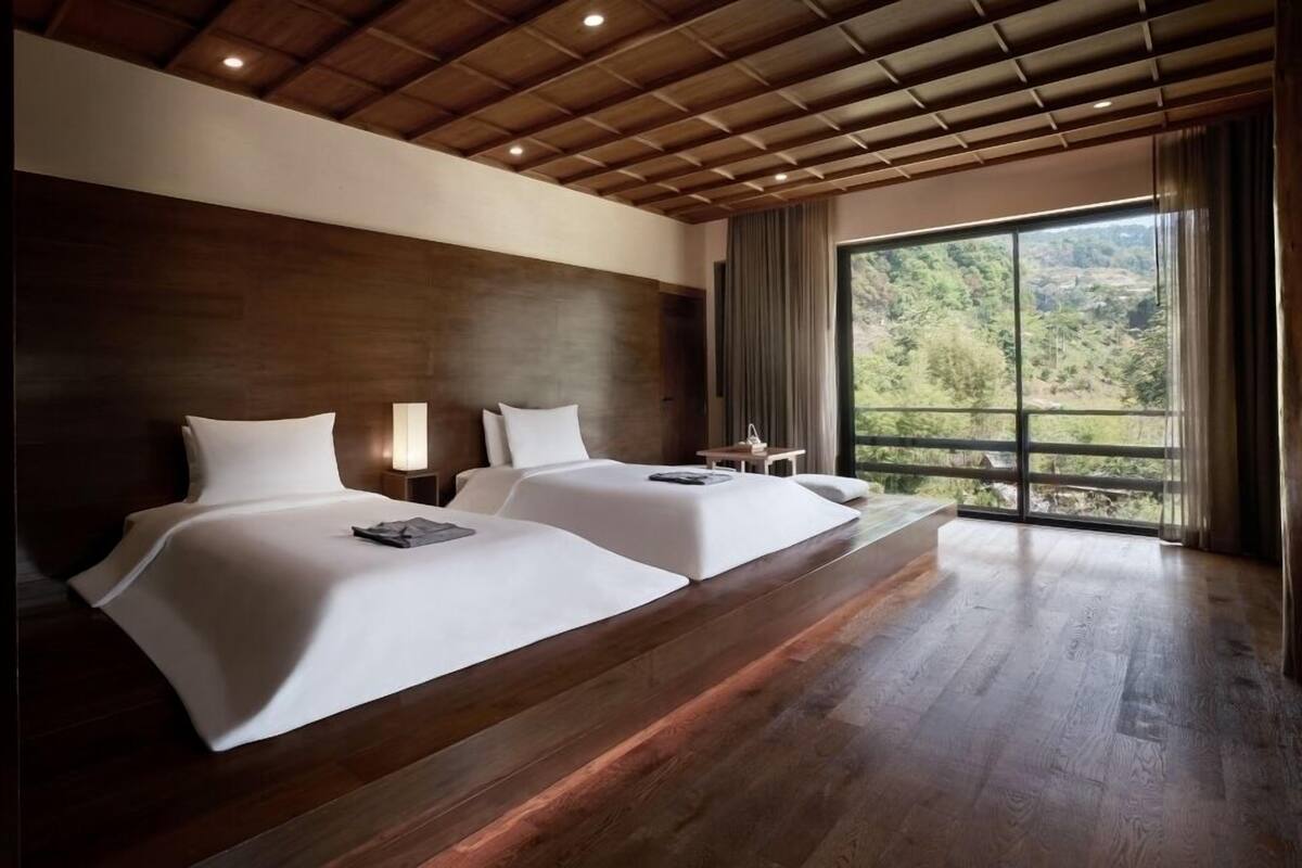 A hotel room with two beds, wooden ceiling, large window with a view of greenery, and a small table with a lamp.