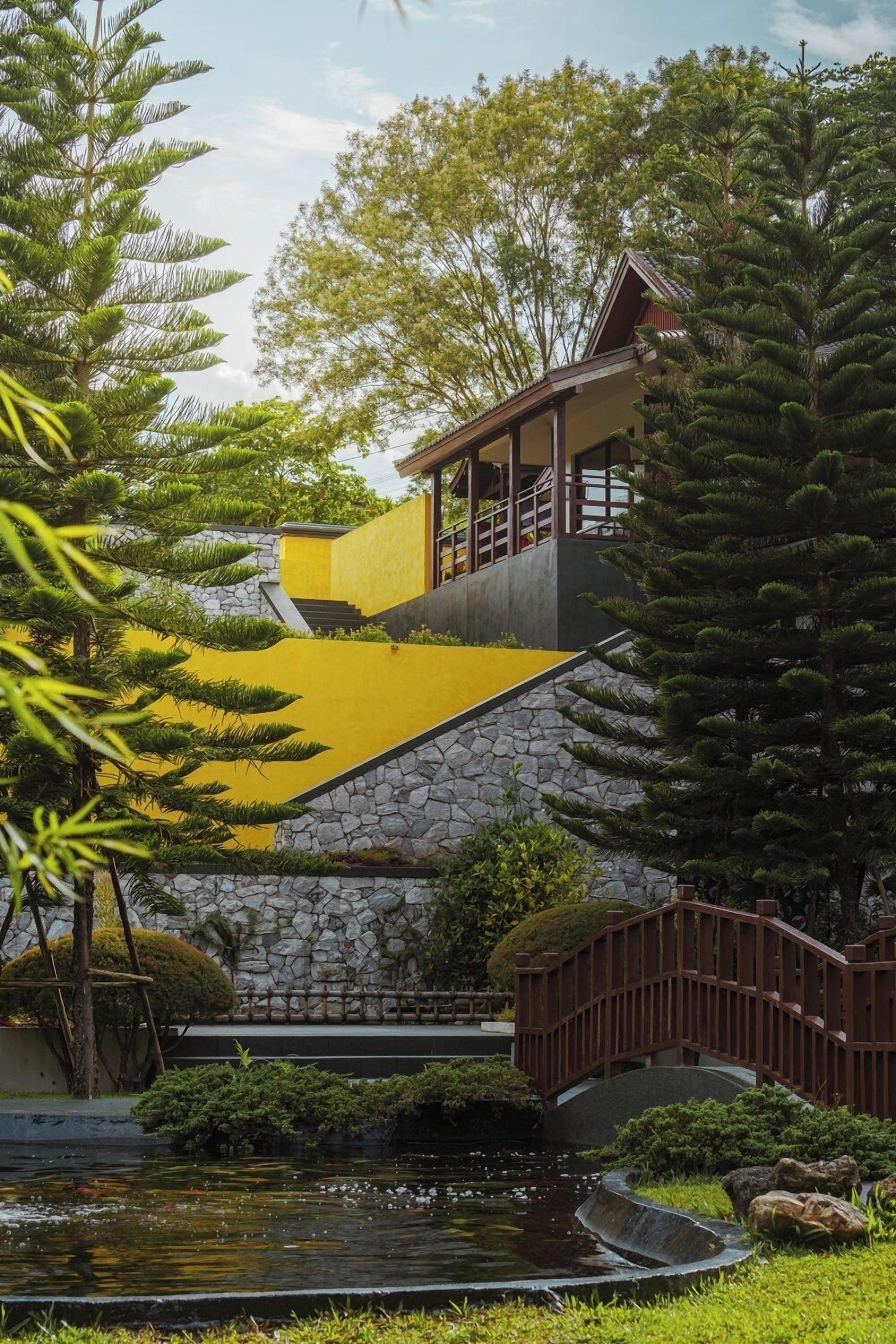 Un bâtiment avec un mur en pierre et une façade jaune, un petit étang et un pont en bois.