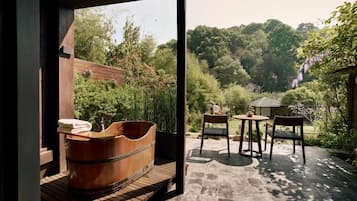 Un baño moderno con una bañera de madera, un banco y un ventanal con vistas a un jardín con mesa y sillas.