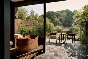 A modern bathroom with a wooden bathtub, a bench, and a window overlooking a garden with a table and chairs.