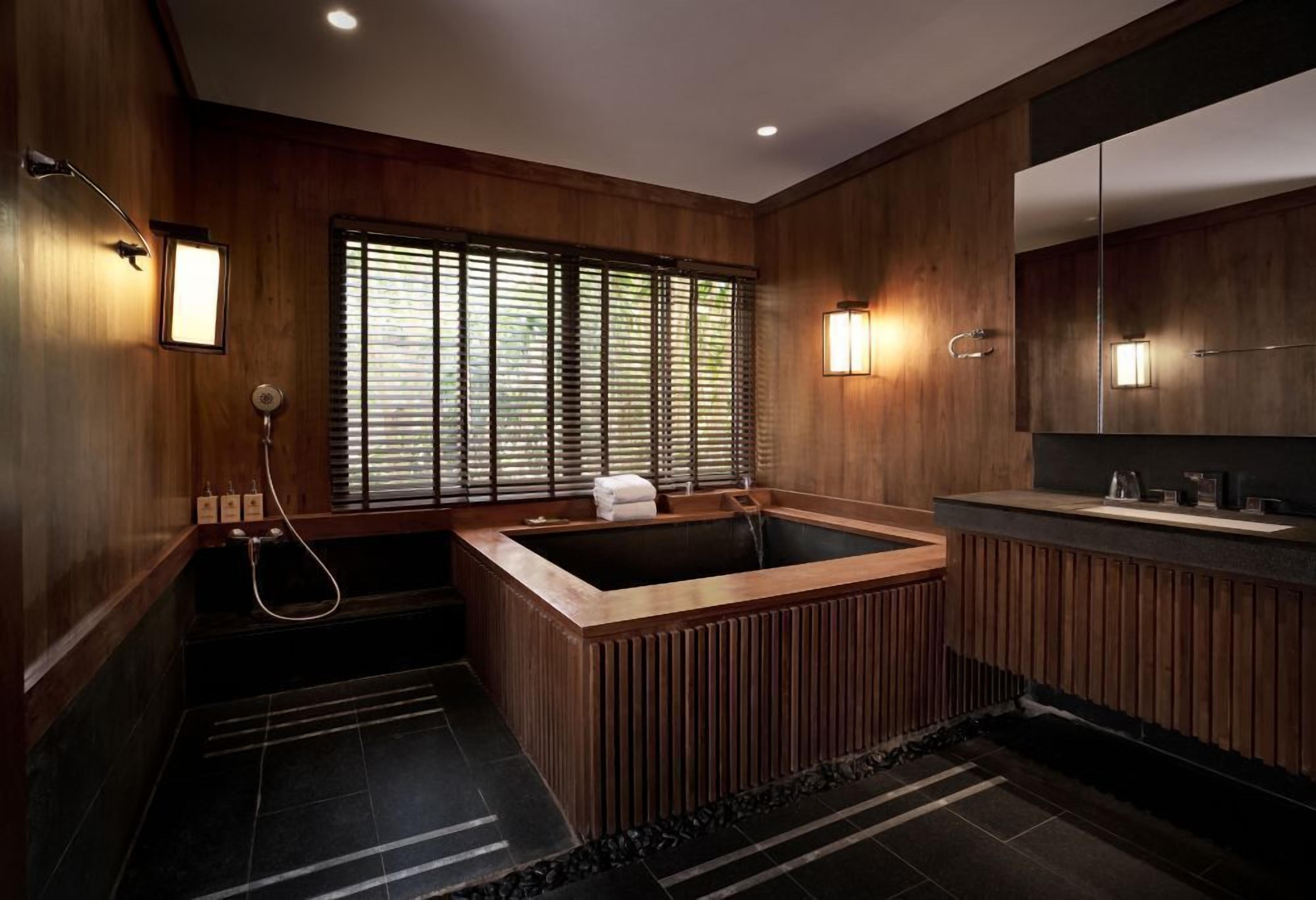A modern bathroom with a wooden bathtub, a large mirror, and a window with blinds.