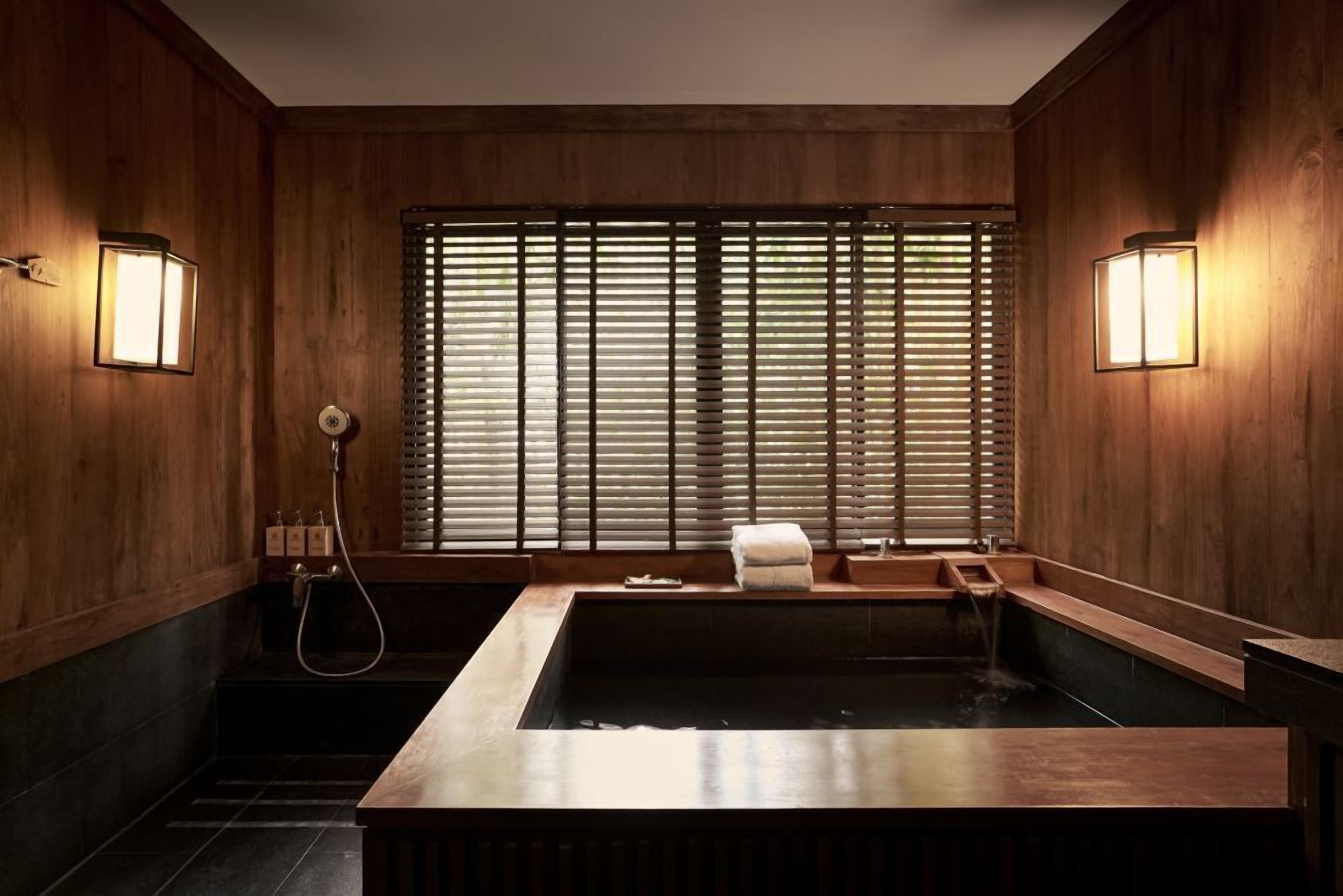 Un interior de madera con una bañera grande, dos luces fijadas a la pared y una ventana con persianas.
