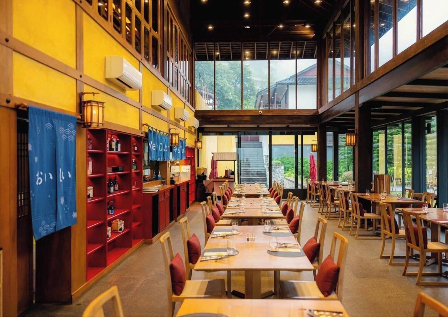 Ein Restaurant mit langen Tischen, roten Regaleinheiten und großen Fenstern mit Blick auf Grünflächen.