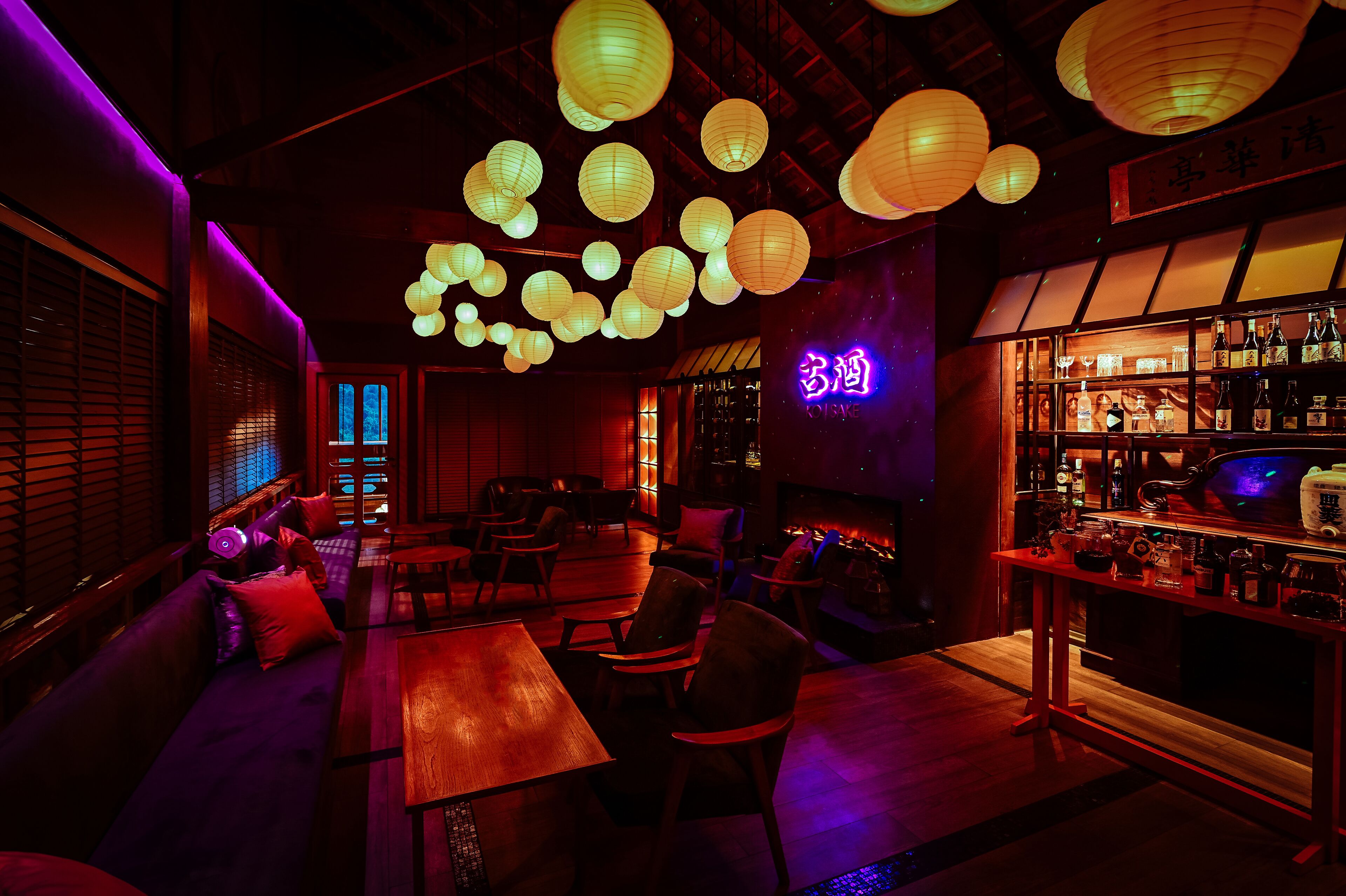 A dimly lit bar area with wooden furniture, hanging paper lanterns, and a neon sign.