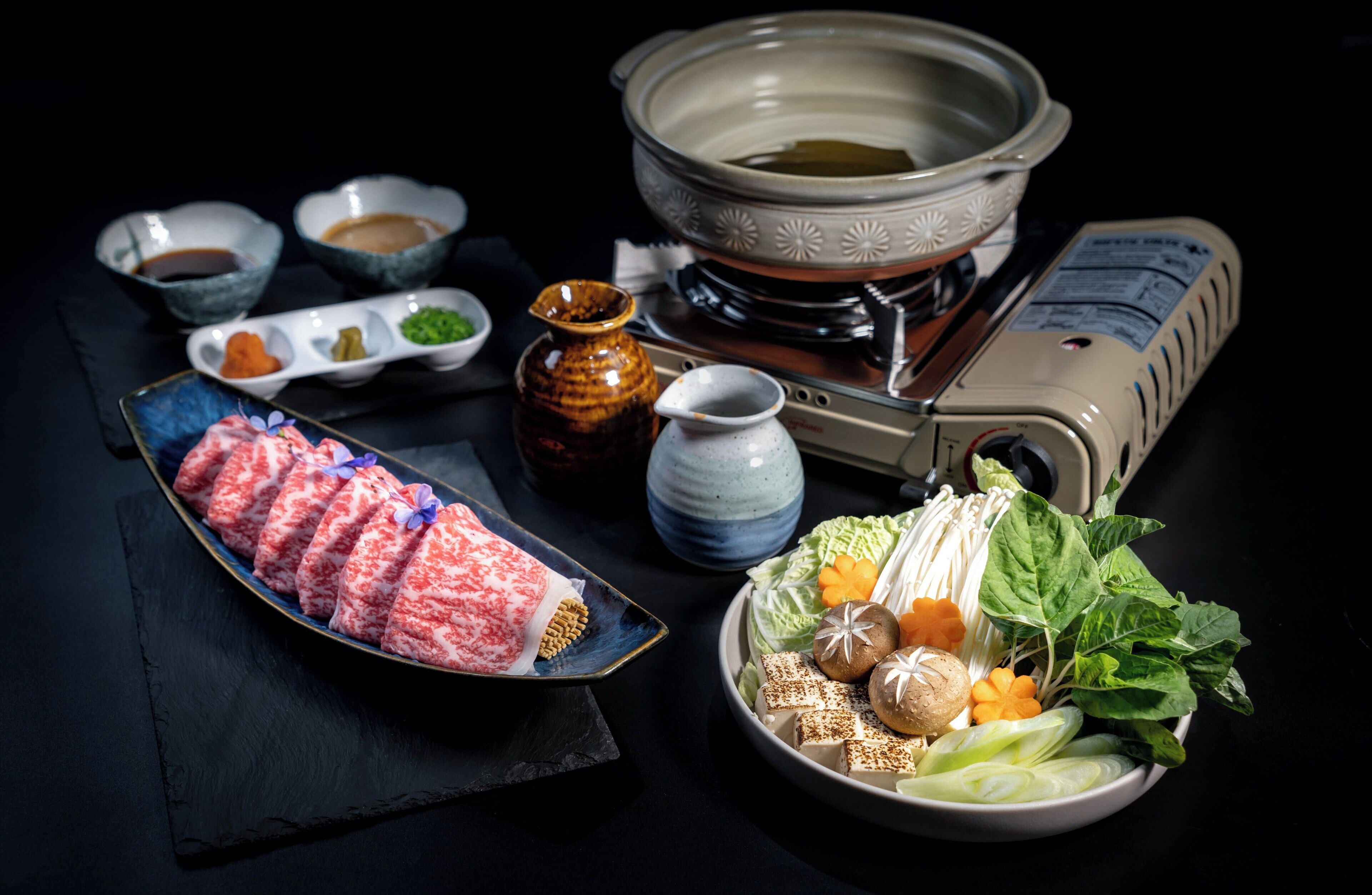A table with a hot pot, various condiments, and a plate of sliced meat.
