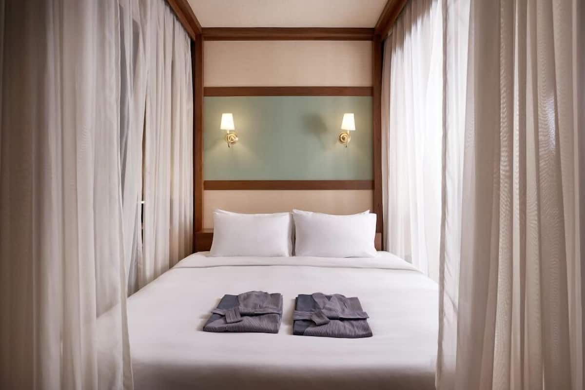 A neatly made bed with two pillows, a headboard with wall-mounted lights, and a pair of folded towels on the bed.