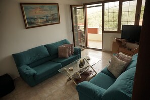 A living room with a teal sofa, a glass coffee table, and a television on a wooden stand. - J&B Family life houset in Agios Matheos (Ag. Mattheos)