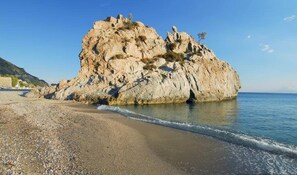 A rocky beach with a large rock formation and calm sea. - Gaea Gardens Studios (Petra-Lesvos)