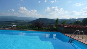 Une piscine d’un bleu éclatant, avec un escalier en métal, entourée d’un mur de pierre et d’une rambarde métallique, d’où l’on peut admirer un paysage montagneux.
