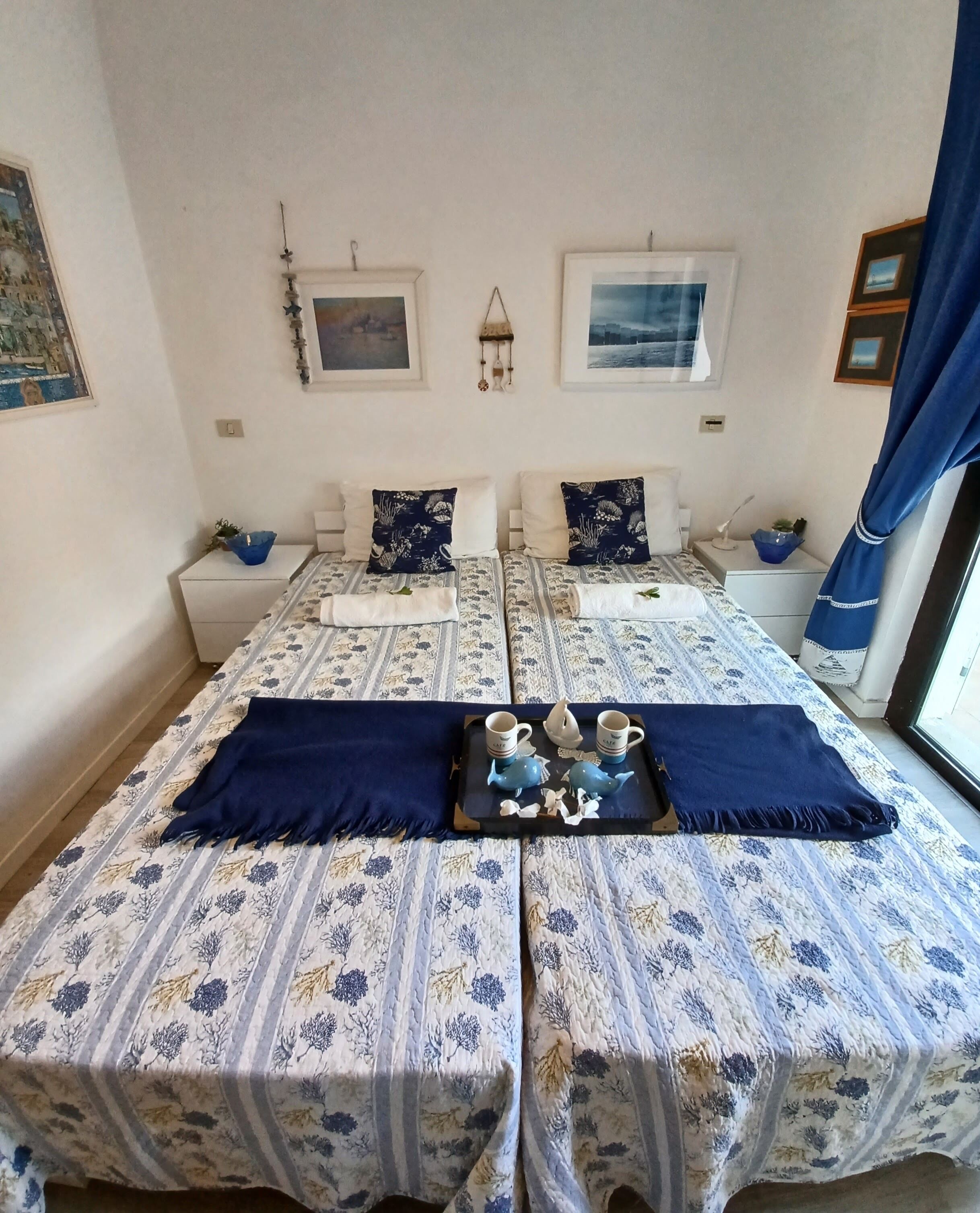 Une chambre double avec deux lits, chacun agrémenté d’une couvre-lit à motifs floraux et d’oreillers assortis. Il y a deux tables de chevet avec des lampes et une petite plante sur chacune. La chambre dispose d’une grande fenêtre avec des rideaux bleus, et des œuvres d’art sont exposées sur les murs.
