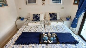 Une chambre double avec deux lits, chacun agrémenté d’une couvre-lit à motifs floraux et d’oreillers assortis. Il y a deux tables de chevet avec des lampes et une petite plante sur chacune. La chambre dispose d’une grande fenêtre avec des rideaux bleus, et des œuvres d’art sont exposées sur les murs.