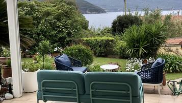 A patio with a green metal sofa and two chairs, overlooking a garden with potted plants and a view of mountains and a lake.