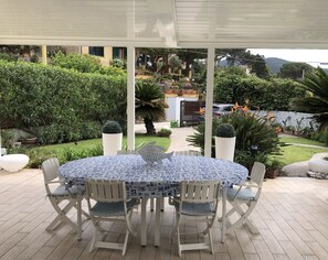 An outdoor patio with a round table and white chairs, surrounded by potted plants and a view of a residential area.