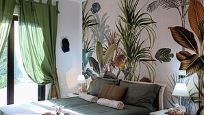 Een slaapkamer met een bed, nachtkastjes, een raam met gordijnen en een wanddecoratie met een tropisch thema.