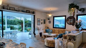 Een moderne woonkamer met een bank, een poef, een eettafel, een keukenhoek en een uitzicht op een tuin door grote ramen.