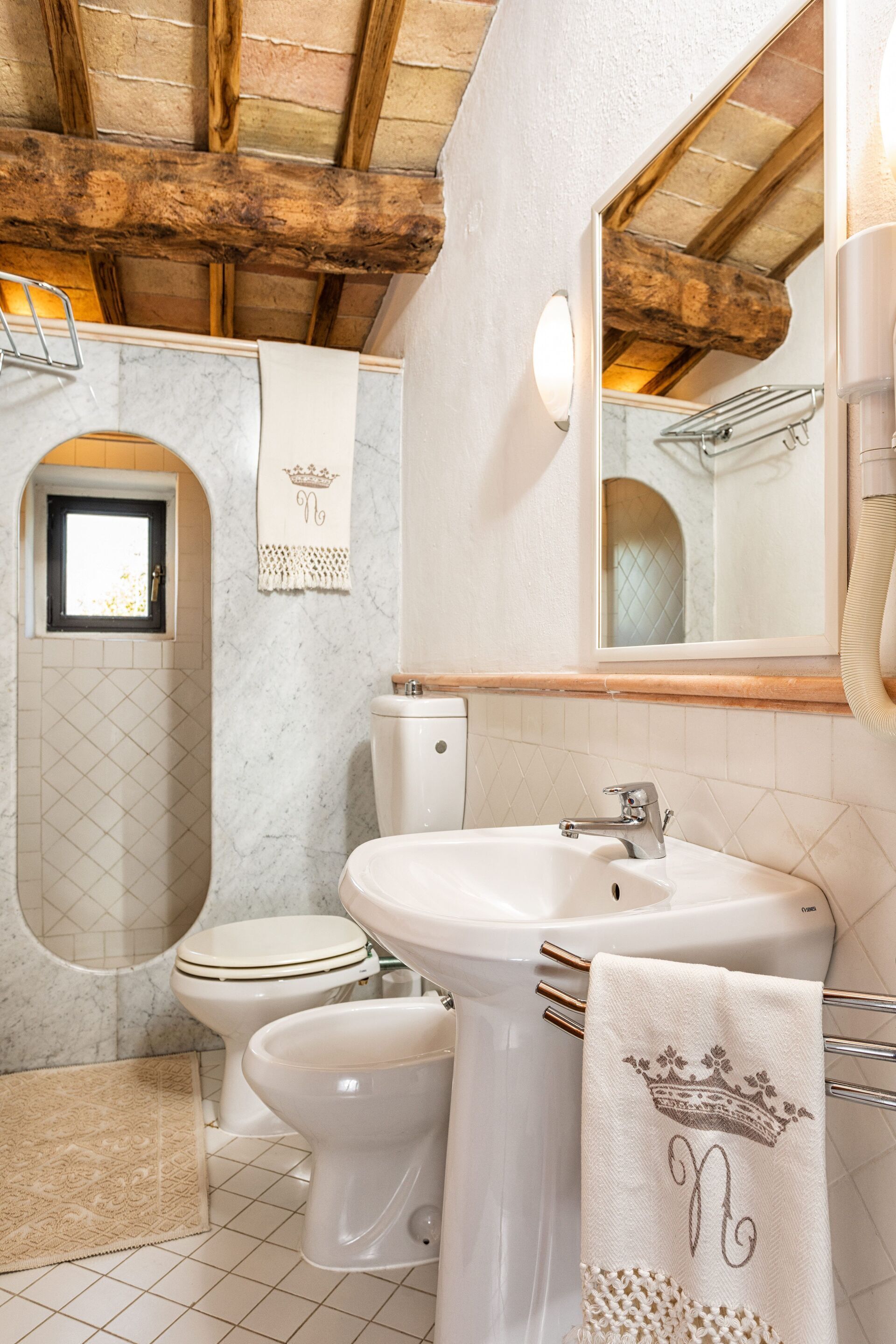 A bathroom with a toilet, bidet, and sink. There is a towel with a monogram hanging on the wall.
