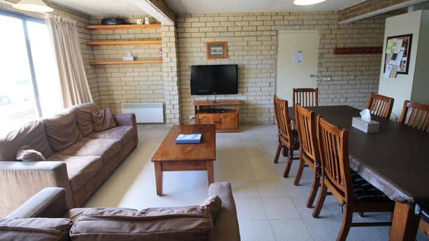 A living room with a brown sofa, a wooden coffee table, and a TV on a stand. There is a brick wall, wooden shelves, and a dining table with chairs. - Central Park 4 - Centre of Jindabyne (Jindabyne)