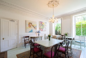 A dining room with a white table, wooden chairs, and a chandelier. There are two paintings on the wall and a window with a view of greenery. - FAMILY & GROUP FRIENDLY - a great location and spacious! (Edinburgh)