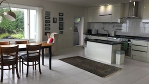 A modern kitchen with white cabinetry, a dining area with wooden chairs, and a view of greenery outside. - Sunday Jeju (Seogwipo)