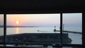 A room with a large window overlooking a sunset over a body of water, a stone wall, and a dock with boats. - Sunday Jeju (Seogwipo)