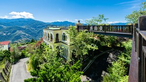 Un edificio de dos pisos con balcón, rodeado de abundante vegetación y con montañas al fondo.