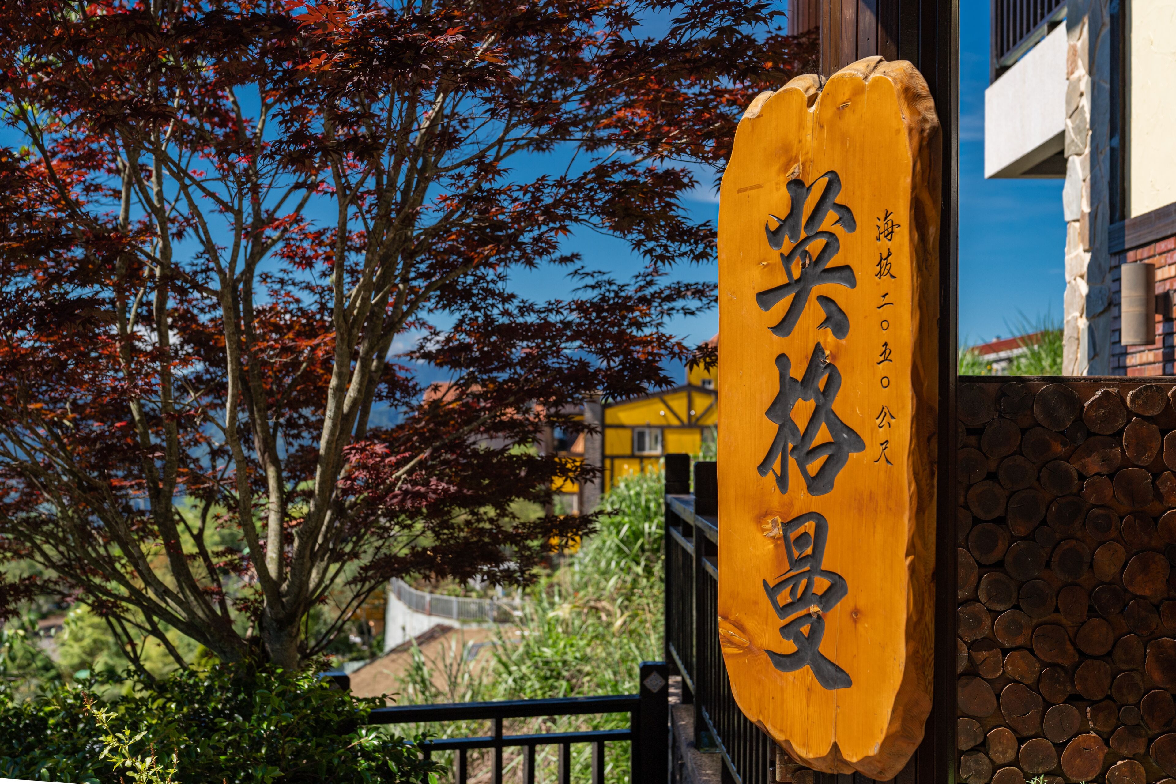 En träskylt med japanska tecken, en stenmur och ett träd med röda löv.