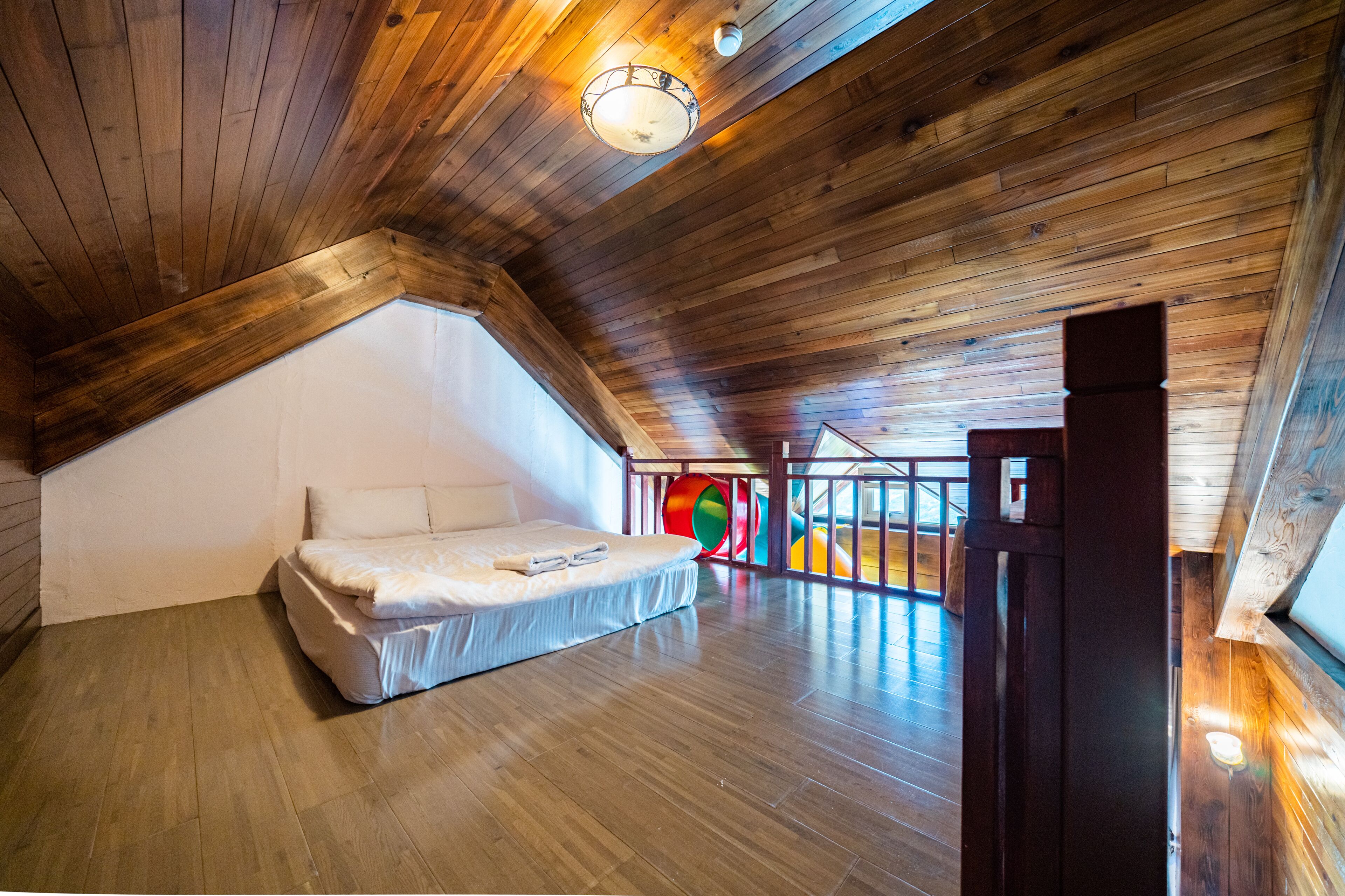 A high-ceilinged attic bedroom with a wooden sloped roof, a bed, and a view of a colorful play area.