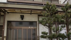 Un edificio japonés tradicional con una puerta de madera grande, un pequeño jardín con un pino y un farol de piedra.