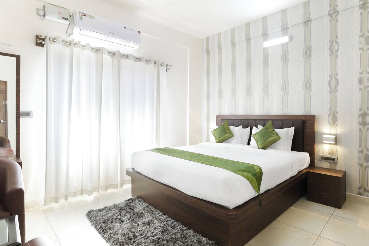 Una habitación de hotel moderna con una cama grande, una mesita de noche con lámpara, zona de ducha y baño con inodoro y lavabo.