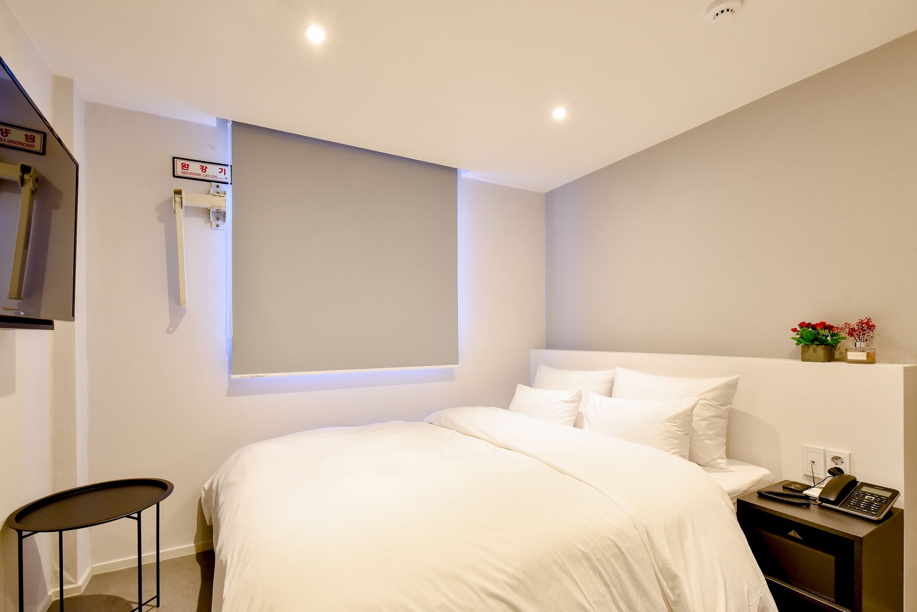 A modern bedroom with a large bed, a small table, a bedside table with a phone, and a wall-mounted shelf with flowers.