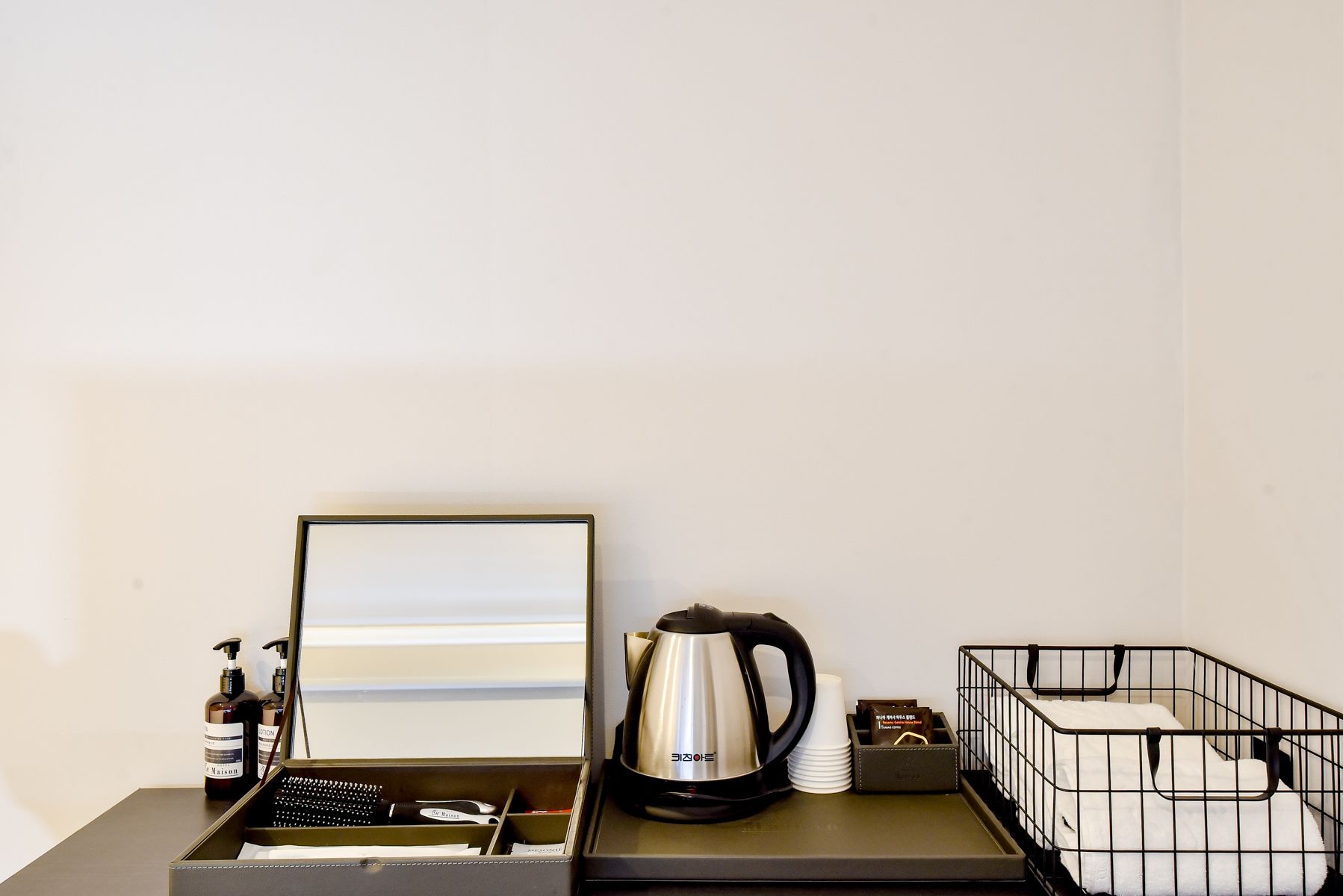 A hotel room with a desk, a kettle, toiletries, a framed picture, and a storage bin.