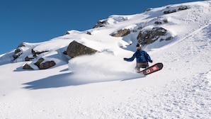 A snowboarder on a snowy slope with rocky terrain and clear skies.