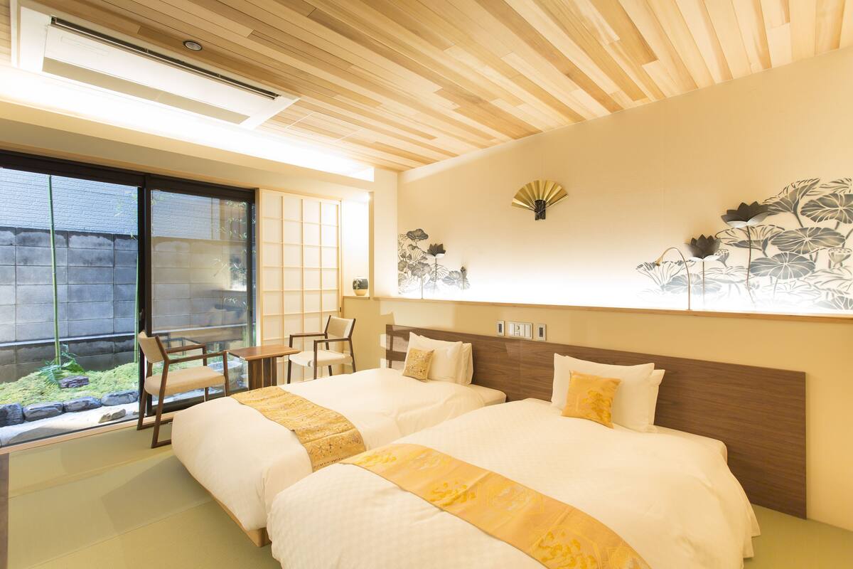 A hotel room with two beds, a wooden ceiling, a large window, and a wall-mounted fan.