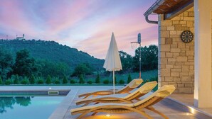 A poolside area with lounge chairs and an umbrella, a stone wall with a dartboard, and a view of a hill with trees. - Gorgeous home in Krnica with sauna (Krnica)