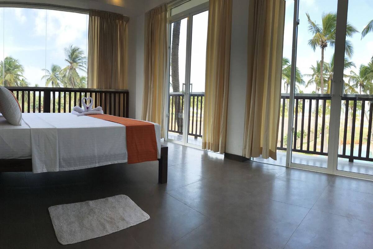 A hotel room with a bed, a nightstand, a lamp, a white rug, and large windows with curtains.