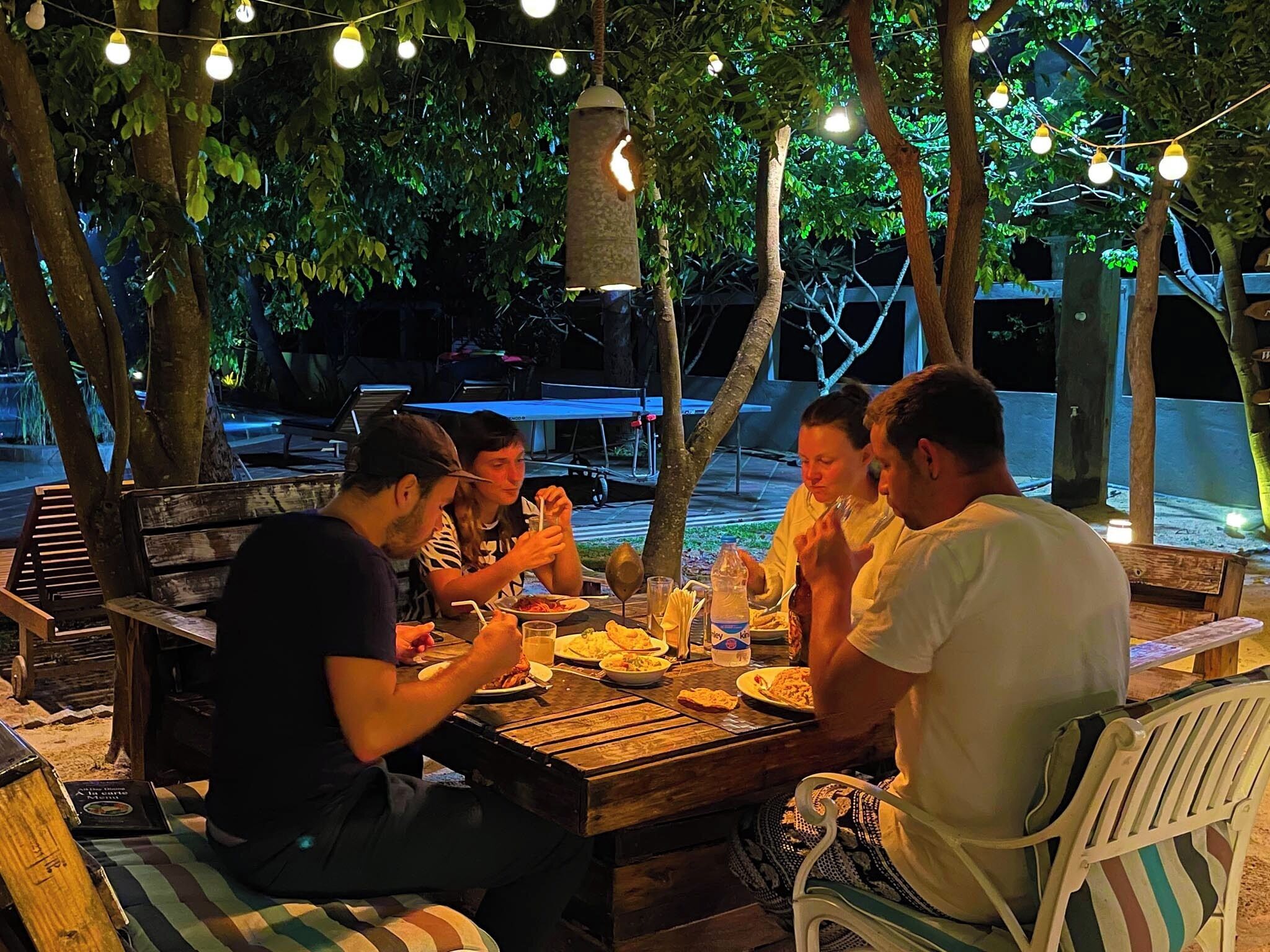 Un grupo de personas cenando en una mesa de madera al aire libre, con luces colgantes sobre ellas, rodeadas de árboles y una estructura cubierta con un toldo azul al fondo.