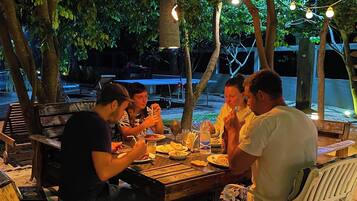 Un grupo de personas cenando en una mesa de madera al aire libre, con luces colgantes sobre ellas, rodeadas de árboles y una estructura cubierta con un toldo azul al fondo.