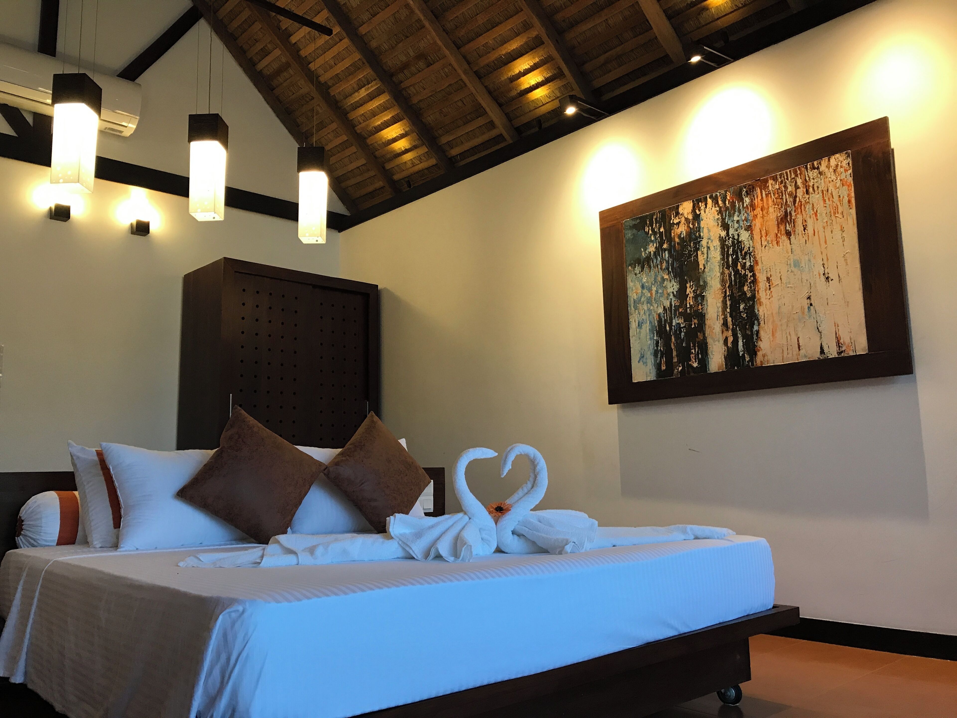 A hotel room with a bed, pillows, a headboard, wall art, and pendant lights.