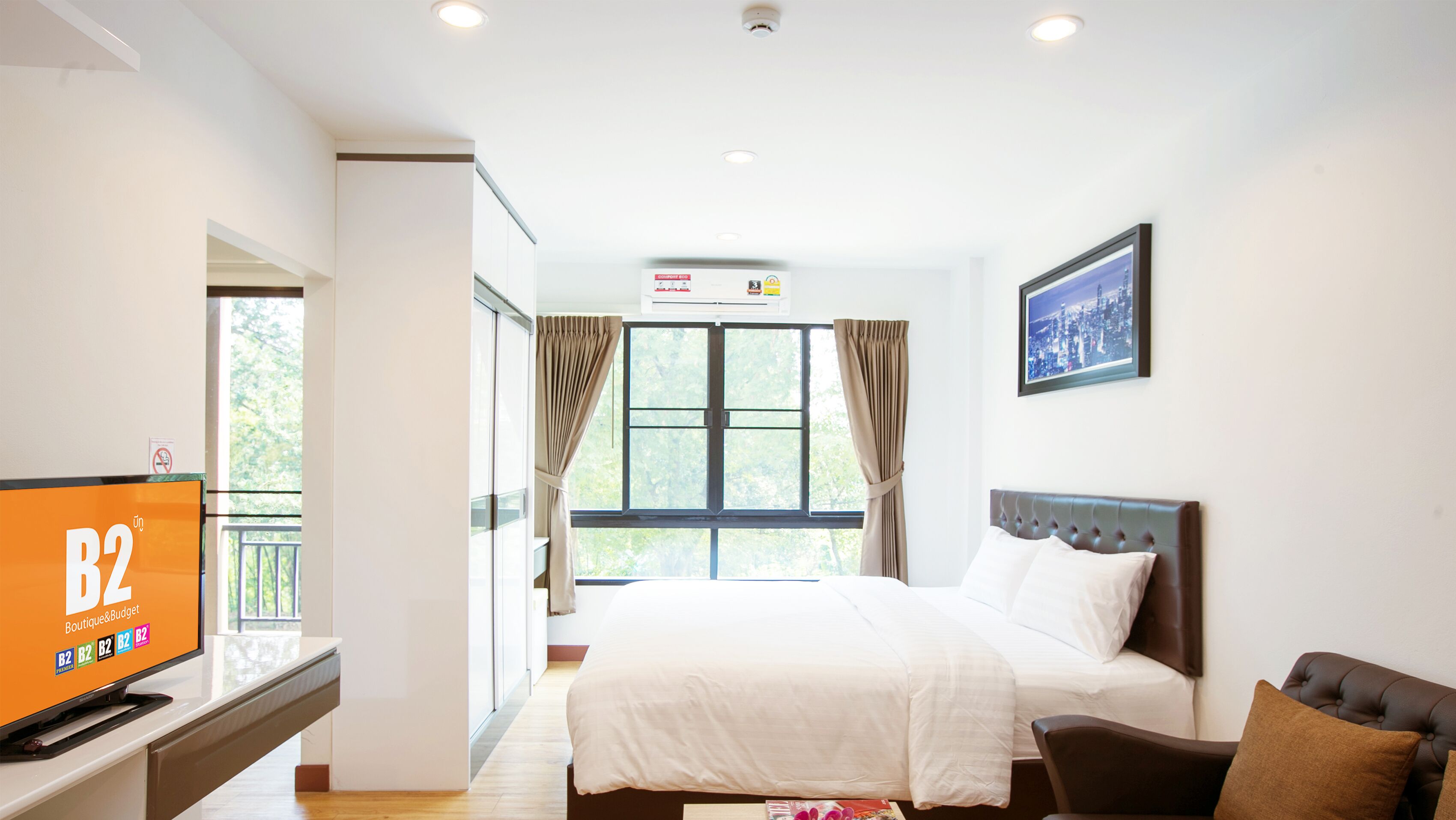 A hotel room with a bed, a TV, a window with curtains, a framed picture on the wall, and an air conditioning unit.